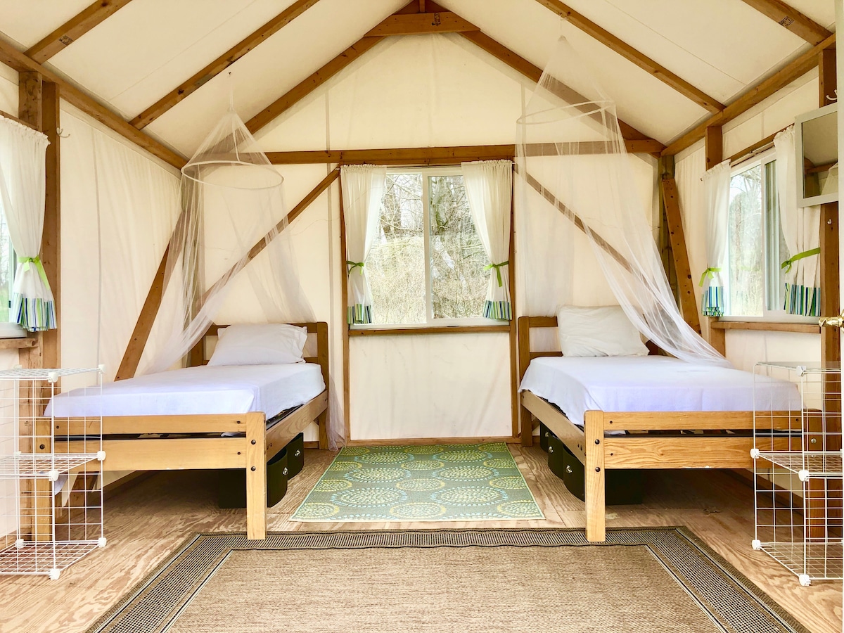 The interior of the cabin features two twin beds placed parallel to each other, each dressed with white linens. Light filters in through screened windows on two sides, enhancing airflow. A patterned rug lies on the wooden floor, contributing to the cozy ambiance.
