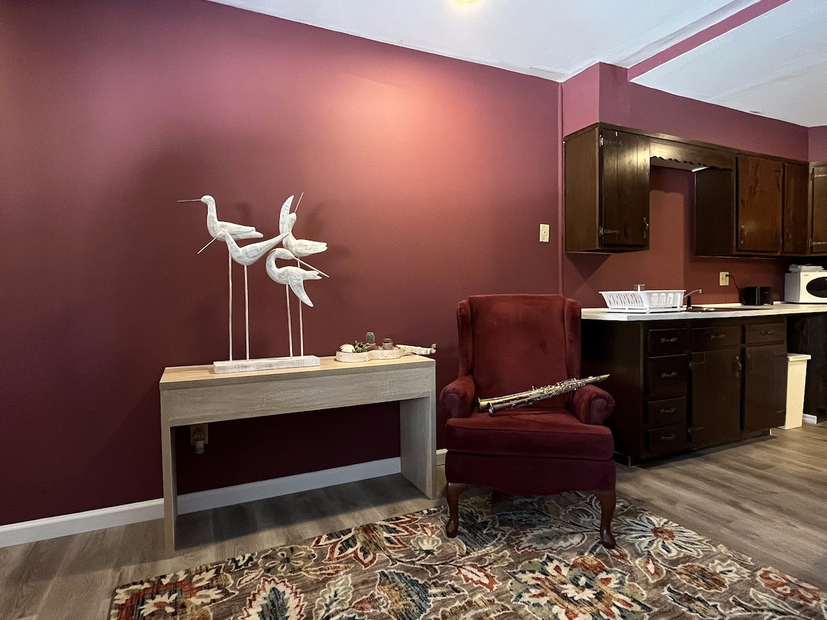 A maroon accent wall complements a wooden console table, which displays decorative bird sculptures. A plush red chair is positioned nearby, adding a touch of comfort. The kitchen area is visible in the background, featuring dark cabinetry and essential appliances.