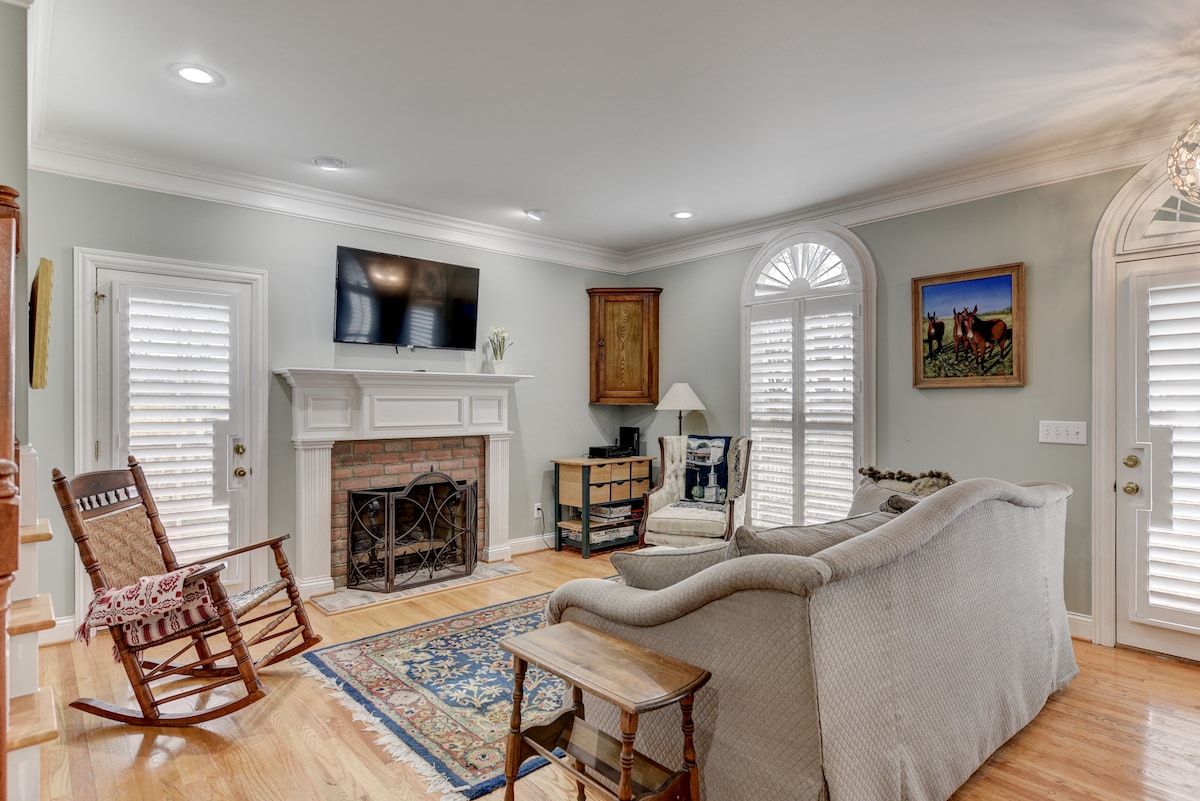 A welcoming living area features a light-colored sofa and a rocking chair beside a wooden side table. The space is illuminated by natural light through arched windows adorned with shutters. A television is mounted above the fireplace, with artwork displayed on adjacent walls.