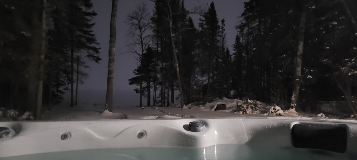 A hot tub is positioned among towering trees in a snowy landscape, offering a serene view of the surrounding nature. The still water reflects the dark night sky, while the soft glimmer of snow adds contrast to the scene.