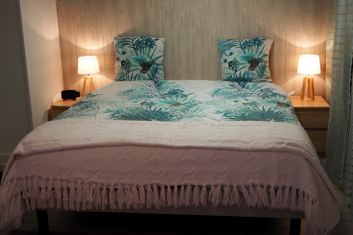 A spacious bed is displayed with a tropical-patterned duvet cover and coordinating cushions. Soft lighting is provided by two bedside lamps, creating a cozy ambiance. A textured blanket with fringed edges is draped over the bed.