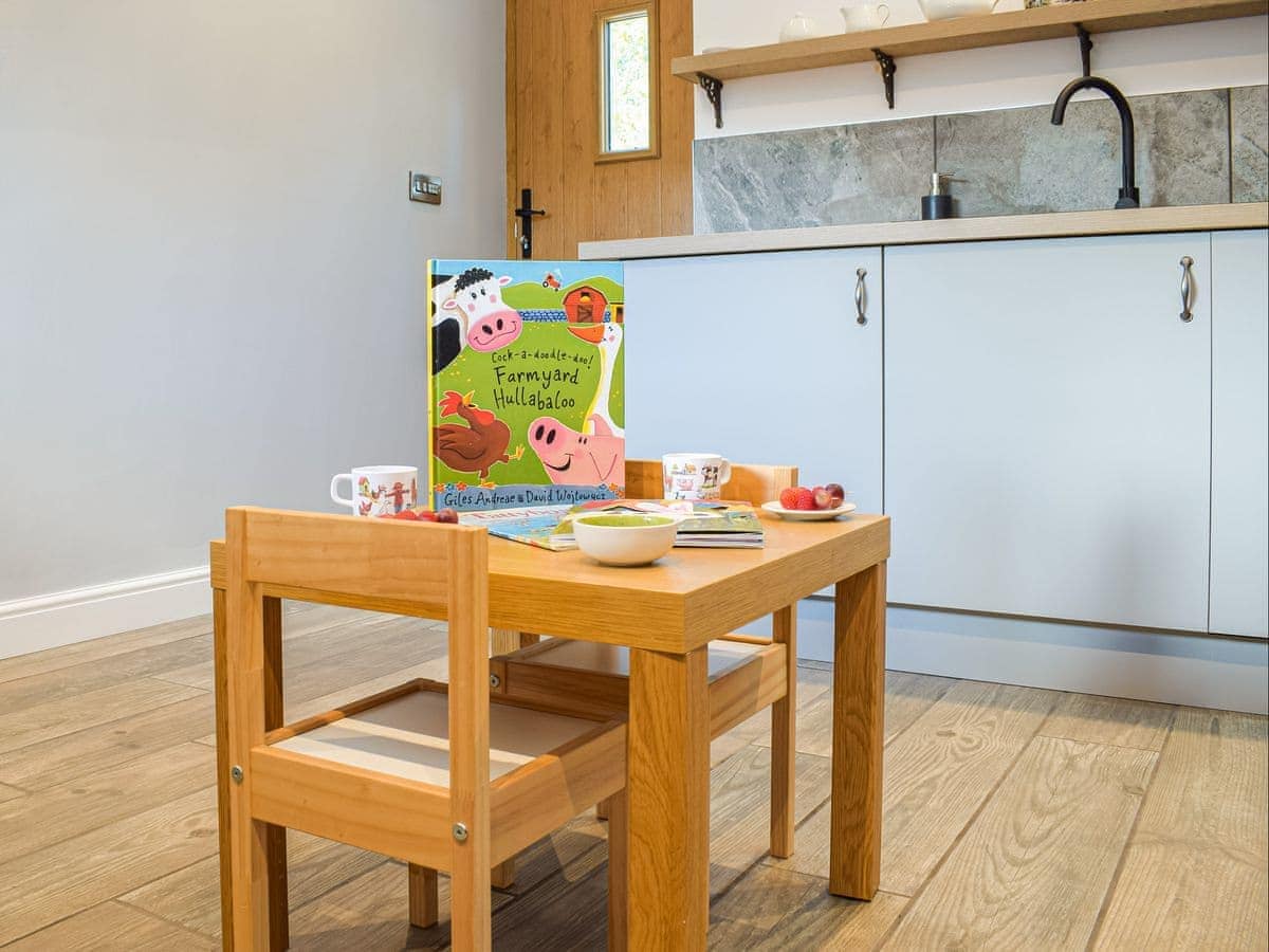 A wooden table is set with children's books, including 'Farmyard Hullabaloo,' and a bowl of apples. Light wooden flooring extends across the area, while modern kitchen cabinetry is visible in the background, featuring pale grey fronts and grey stone countertops.