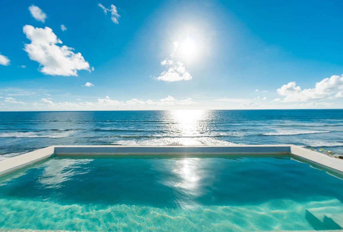 An infinity pool extends towards the horizon, blending seamlessly with the azure sea. The sun reflects off the water's surface, creating a serene and calming view. Fluffy white clouds drift across a bright, open sky, enhancing the tranquil atmosphere.