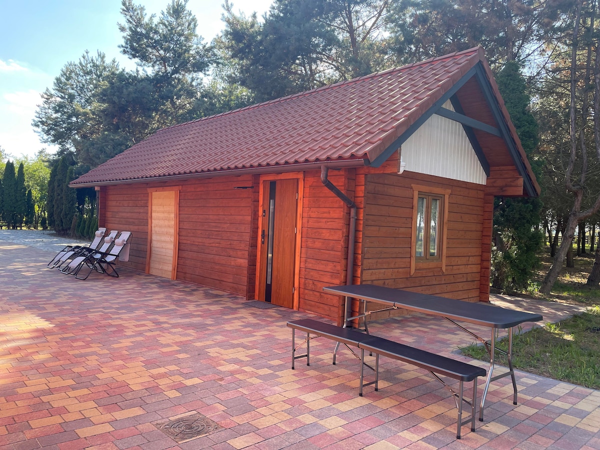 A wooden cabin is shown, featuring a sloping roof and natural wood façade. A small porch entry is visible, flanked by a wooden bench and a folding table with seating. Sunlight illuminates the scene, highlighting the surrounding greenery.