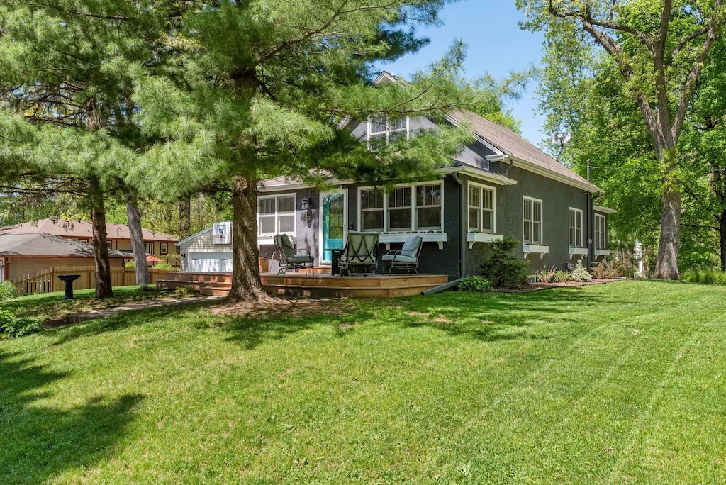 A charming house sits among lush greenery, featuring large windows that allow natural light to fill the interior. A wooden deck extends from the front, offering seating areas in a serene outdoor space surrounded by trees.