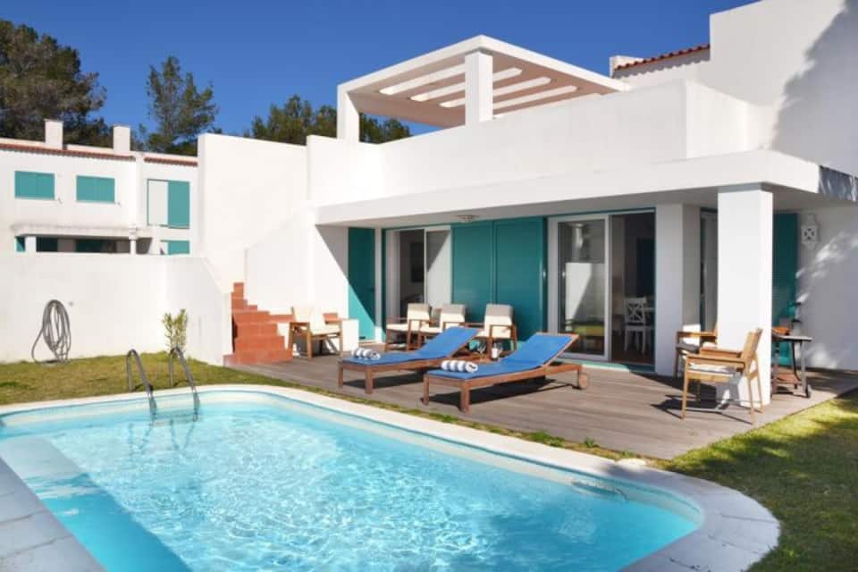 A two-story beach villa is surrounded by a well-maintained lawn. A private swimming pool is visible, complemented by wooden loungers and umbrellas. The exterior features large glass doors that open to a bright, welcoming outdoor area. Sunlight enhances the clean, modern architectural design.
