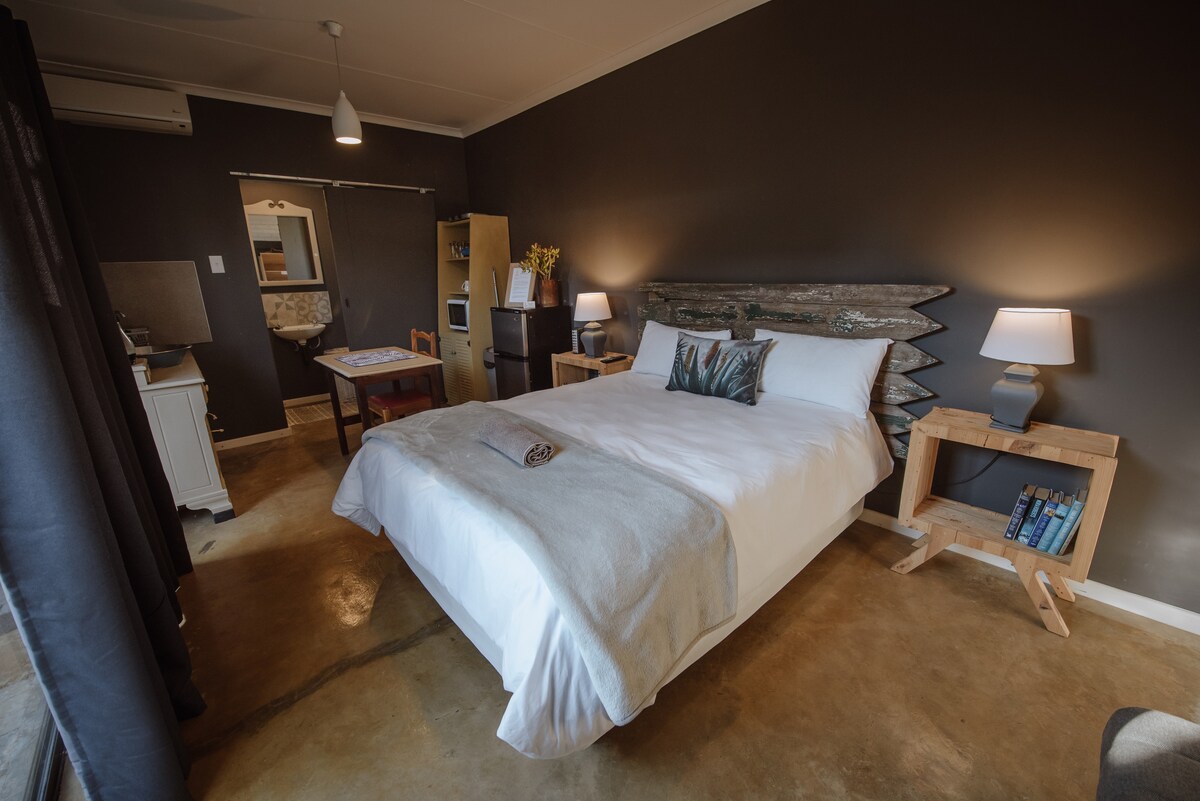 A spacious sleeping area features a queen-sized bed with a textured headboard and soft bedding. Two wooden bedside tables, each with a lamp, provide illumination. Natural light enters through large curtains, revealing a cozy corner with a small table and chairs.
