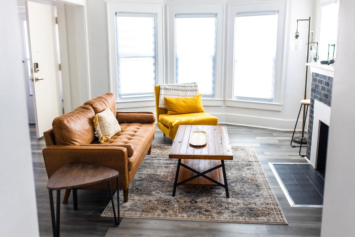 The living room showcases a warm brown leather sofa and a vibrant yellow accent chair, complemented by a wooden coffee table centered on a decorative area rug. Natural light streams through three large windows, illuminating the space with a bright, inviting ambiance.