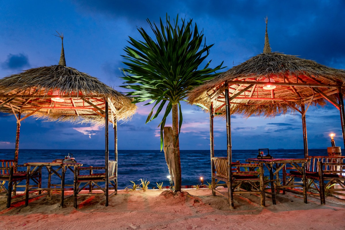 Two traditional beach huts with thatched roofs are situated on sandy shores, facing the ocean at twilight. Wooden tables and chairs are arranged under the huts, and a palm tree is centrally located. Soft lighting enhances the serene setting as the waves gently lap the beach.