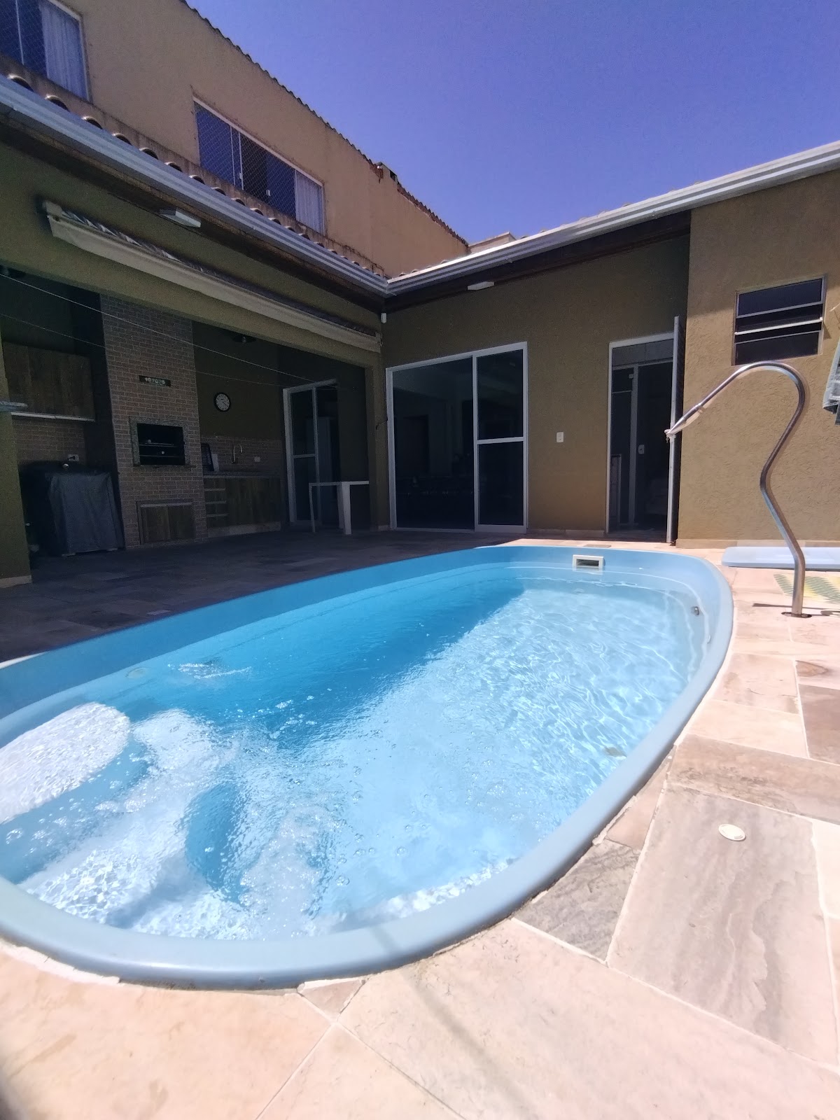 A private pool is visible, featuring clear blue water and a gentle ripple on the surface. Surrounding the pool, a tiled patio provides ample space for relaxation. Glass doors and windows can be seen in the background, leading into a shaded outdoor area.