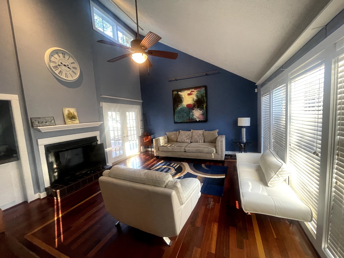 A spacious living area is highlighted by high vaulted ceilings and large windows allowing natural light to fill the room. Comfortable seating is arranged around a central rug, with a modern clock and a painting accenting the walls. A sleek entertainment unit is positioned beneath the wall-mounted television.