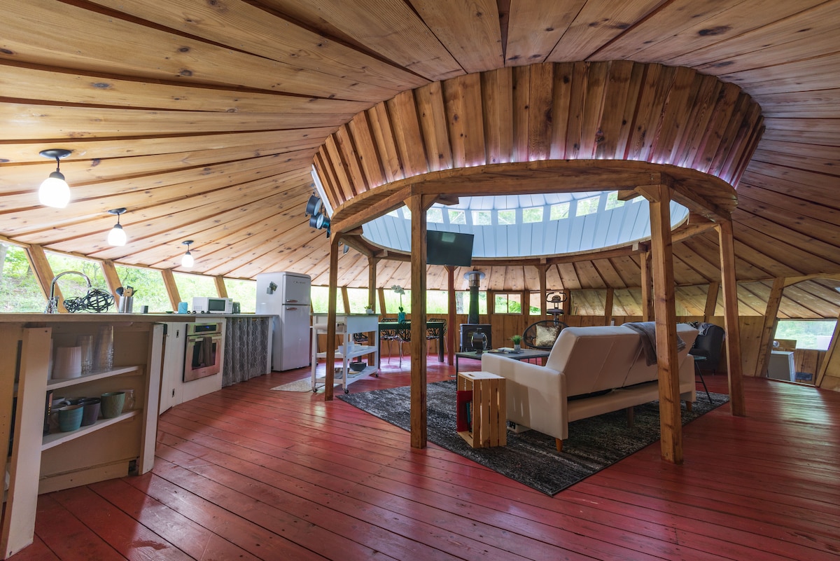 The interior of the wooden round house features a spacious open concept with wooden ceilings and red flooring. A white sofa is positioned on a textured rug. The kitchen area includes modern appliances, with natural light streaming through numerous windows along the curved walls.
