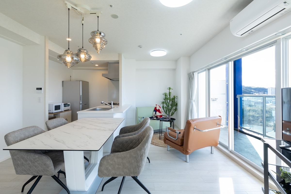 A bright dining area features a sleek white table surrounded by four upholstered chairs, complemented by pendant lighting. A comfortable sofa sits nearby, while large windows invite natural light and provide views of the surrounding area. A small kitchen area is visible in the background.