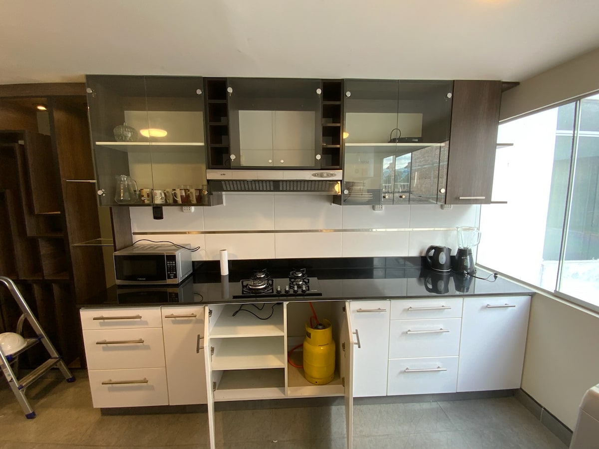 A modern kitchen is equipped with a combination of dark wood and white cabinetry, featuring a sleek black countertop. Appliances include a microwave, blender, and electric kettle, arranged neatly beside a gas stove. Natural light fills the space through a window, enhancing the practical layout.