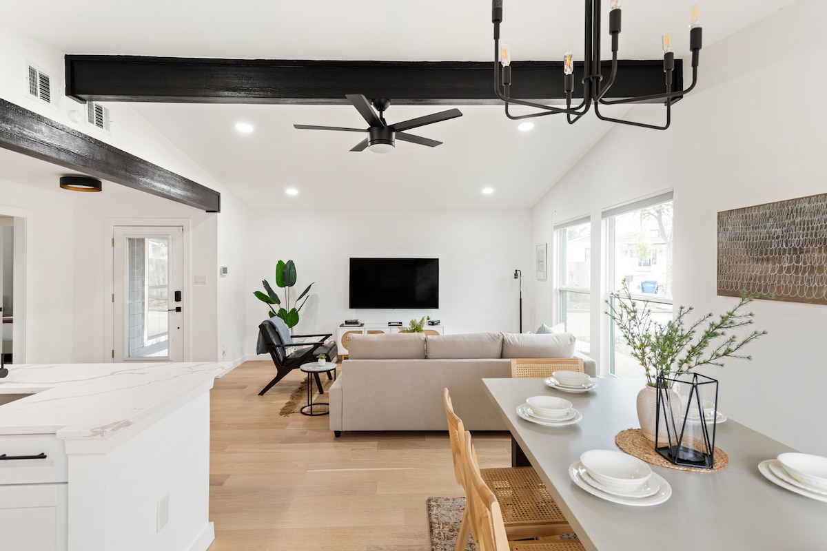 A spacious living area is displayed with a large beige sofa facing a flat-screen television mounted on the wall. Natural light floods the room through multiple windows, illuminating a dining table set for four. A stylish ceiling fan and modern decor complete the inviting atmosphere.