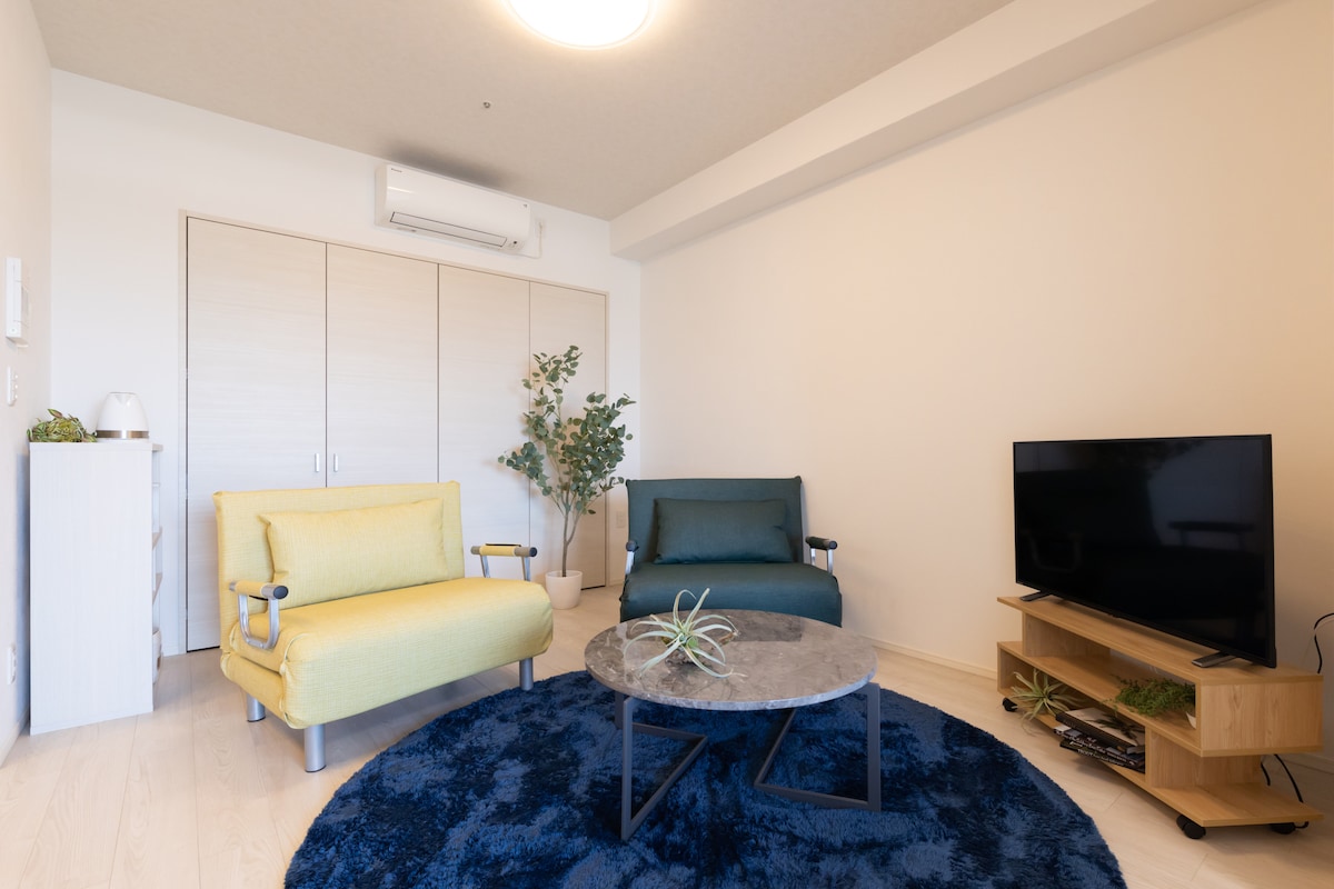 A cozy living area is presented with a yellow armchair and a teal sofa, arranged around a circular coffee table. A soft blue rug covers the floor, while a television is situated on a wooden stand. Indoor plants add a touch of nature to the space.