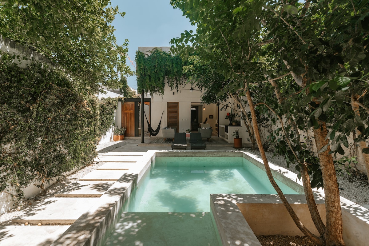 A tranquil outdoor area features a swimming pool surrounded by lush greenery and natural stone. Comfortable seating options are placed nearby, along with a traditional hammock, providing a relaxed atmosphere for enjoying the outdoor space.