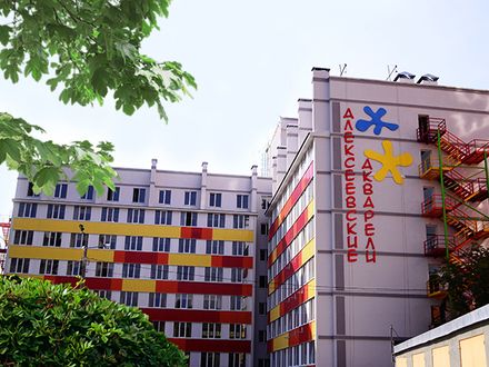 A colorful multi-story building features a blend of red, yellow, and blue stripes. The front displays decorative motifs and the name 'Алексевские акварели' prominently. Lush green foliage provides a natural frame, enhancing the vibrant architecture of the property.