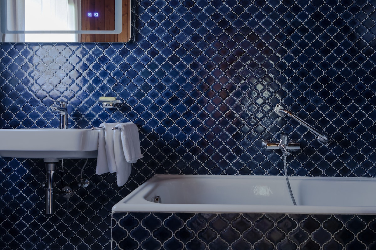The bathroom features dark blue hexagonal tiles that create a bold visual effect. A white bathtub and a wall-mounted sink are present, accompanied by neatly hung white towels. Light reflects off the tiles and a mirror above the sink adds to the space's functionality.