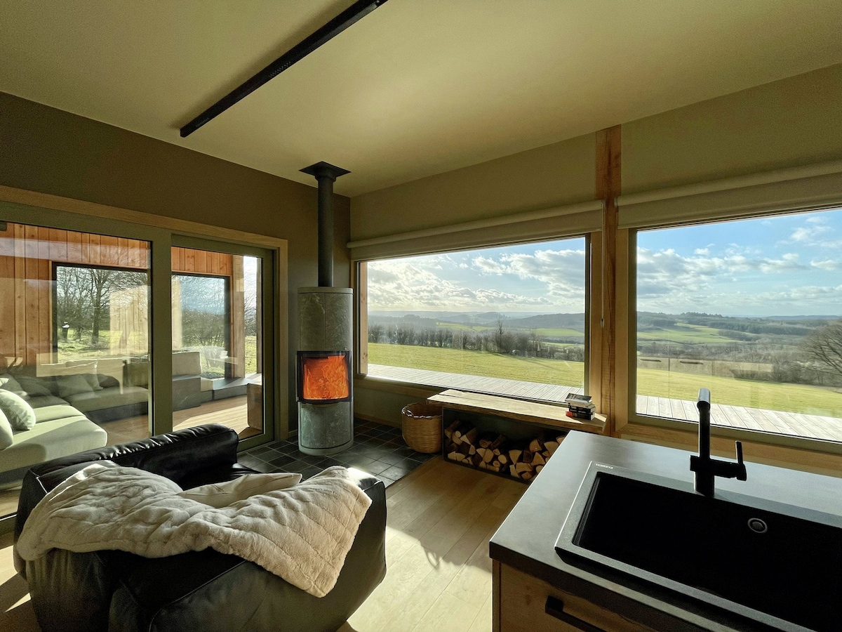 A cozy living area features large windows that provide a panoramic view of the surrounding prairies and woods. A wood-burning stove is positioned for warmth, while a stack of firewood sits nearby. A sink is visible in the foreground, enhancing the open layout.