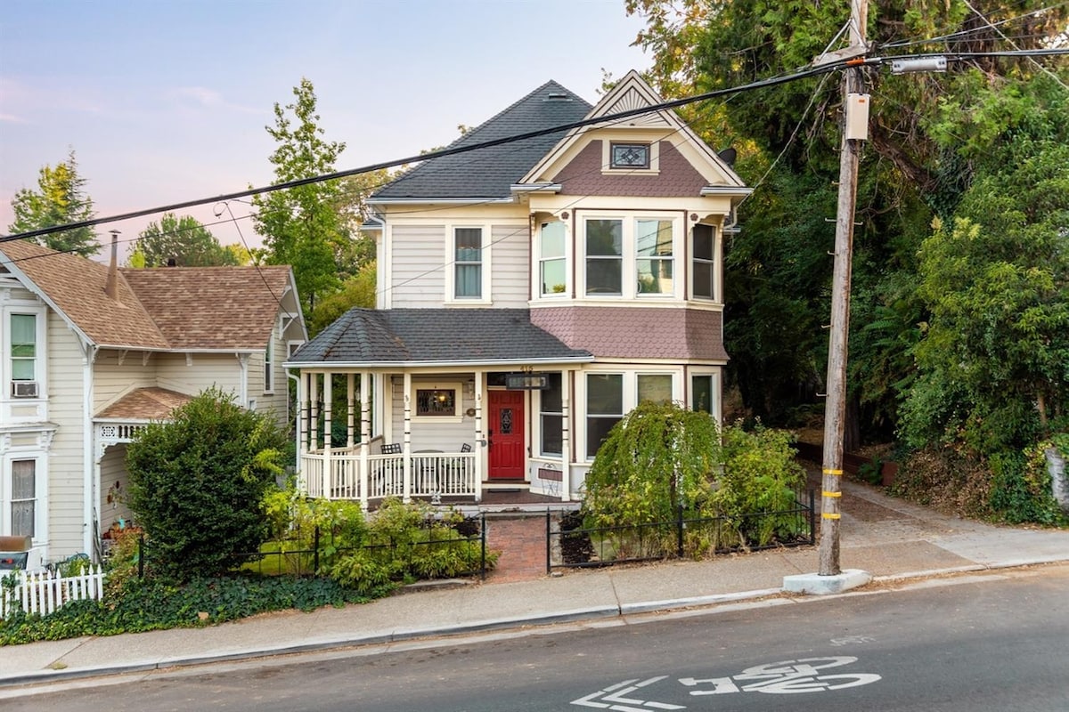 An elegant two-story Victorian home is depicted, featuring a wraparound porch with white railings and a striking red front door. Lush greenery and carefully maintained landscaping border the property. The house is set against a peaceful residential street framed by trees, with a clear sky above.