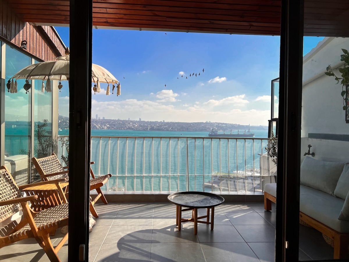 A serene balcony space offers a panoramic view of the shimmering water and distant cityscape. Wooden furniture, including a sofa and two chairs, complement the outdoor setting. A cantilever patio umbrella provides shade, while the inviting atmosphere invites relaxation.