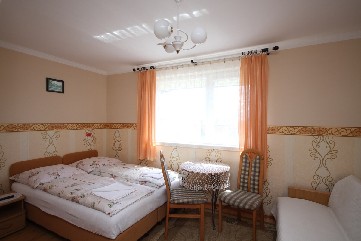 A well-lit bedroom features a double bed with floral bedding, complemented by soft natural light from the large window. A small round table with two cushioned chairs occupies one corner, while a cozy sofa adds additional seating along the wall.