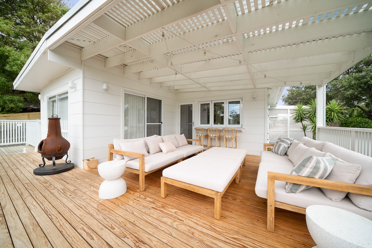 An outdoor living space features wooden decking with a spacious lounge area, complemented by light-colored cushions. A communal dining area with bar stools is situated adjacent to large windows. A sitting area has a modern fire pit, and lush greenery frames the space.