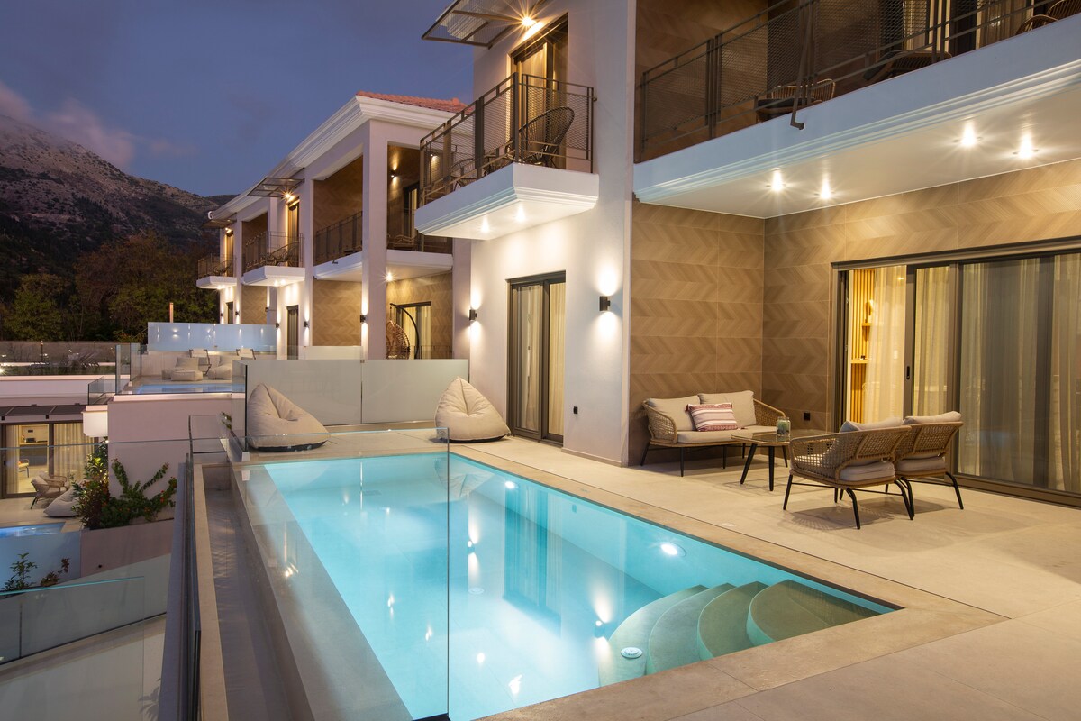 An outdoor area features a modern pool surrounded by inviting loungers and seating. Soft lighting enhances the space, while a clear view of the mountains creates a serene backdrop. Multiple levels of the building are visible, showcasing balconies with seating arrangements.
