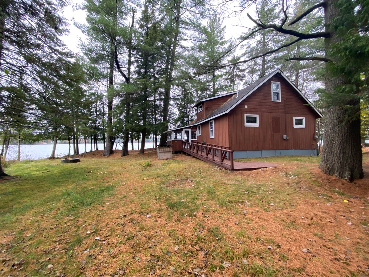 The cabin, constructed in a rustic style, is framed by towering trees and overlooks Raquette Lake. A spacious deck extends from the building, offering a spot for relaxation. A fire pit is visible in the grassy yard, surrounded by a carpet of pine needles.