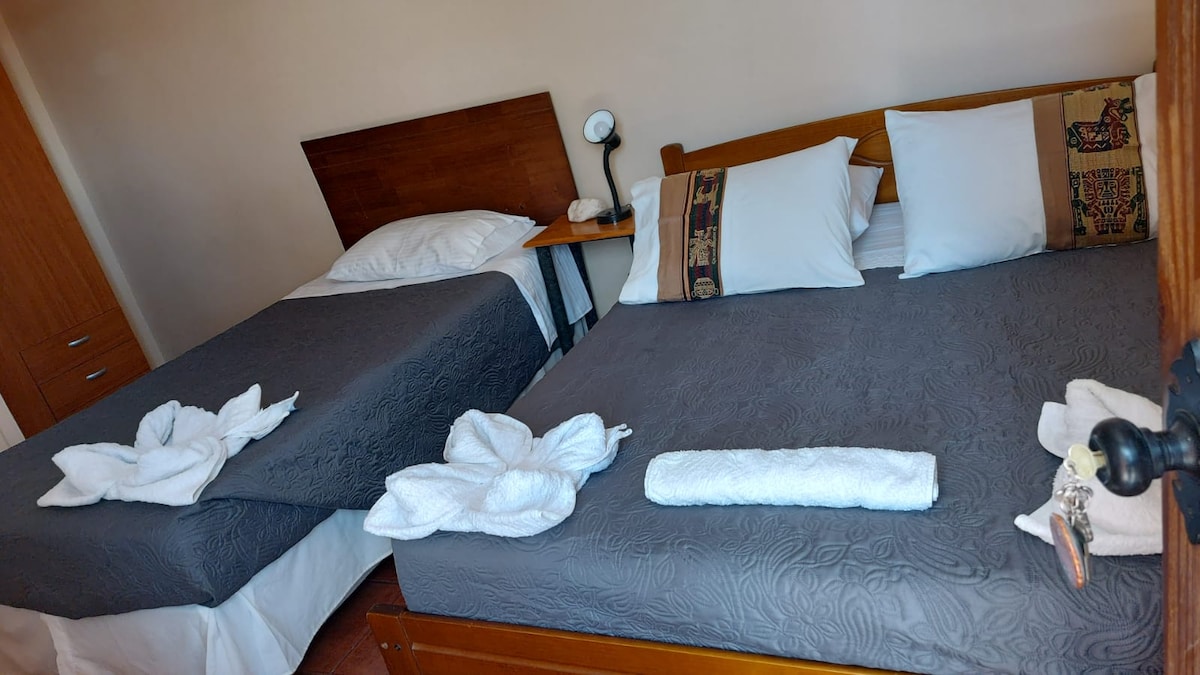 The room features two spacious beds with soft linens and decorative pillows. A bedside lamp is positioned on a wooden nightstand alongside each bed. A set of neatly arranged towels is placed on the bedding, contributing to a tidy appearance.