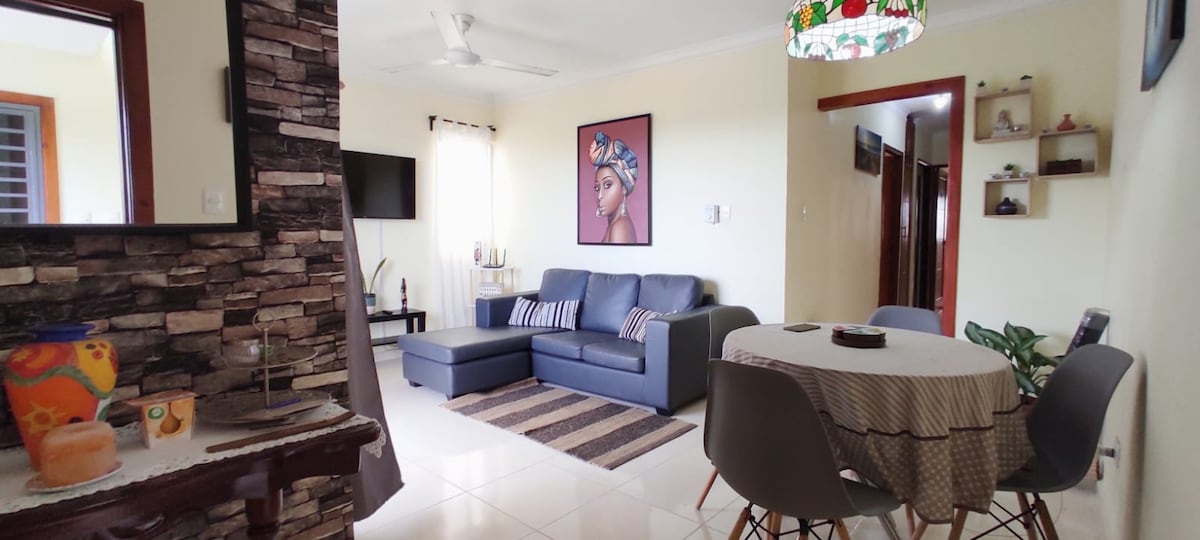 A spacious living area features a modern sectional sofa and a circular dining table with stylish chairs. Natural light enters through a window draped with light curtains, highlighting the neutral color palette and decorative accents on the walls.
