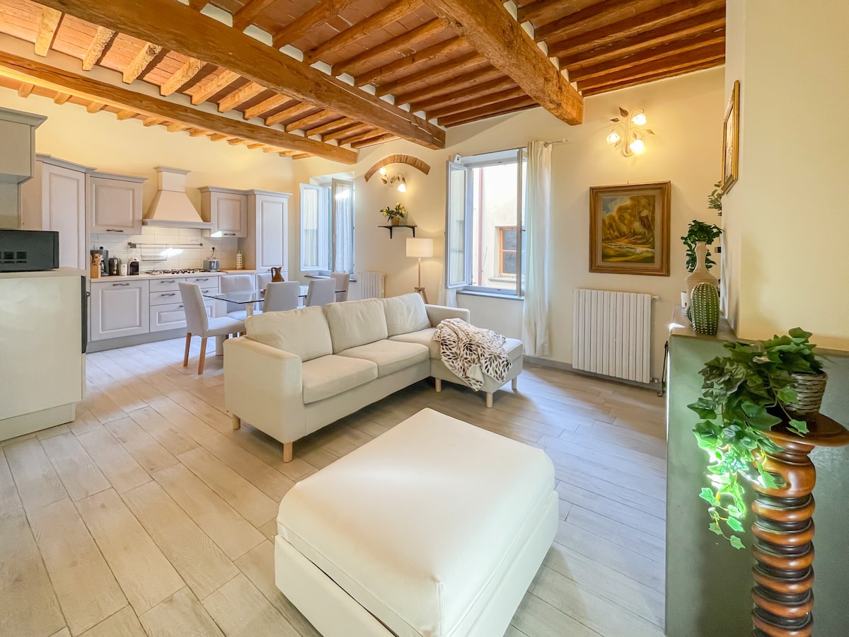 A spacious living area features a light-colored sofa with a decorative throw, complemented by wooden beams overhead. The open kitchen with modern appliances is visible in the background, alongside large windows that provide natural light and a view of the exterior.