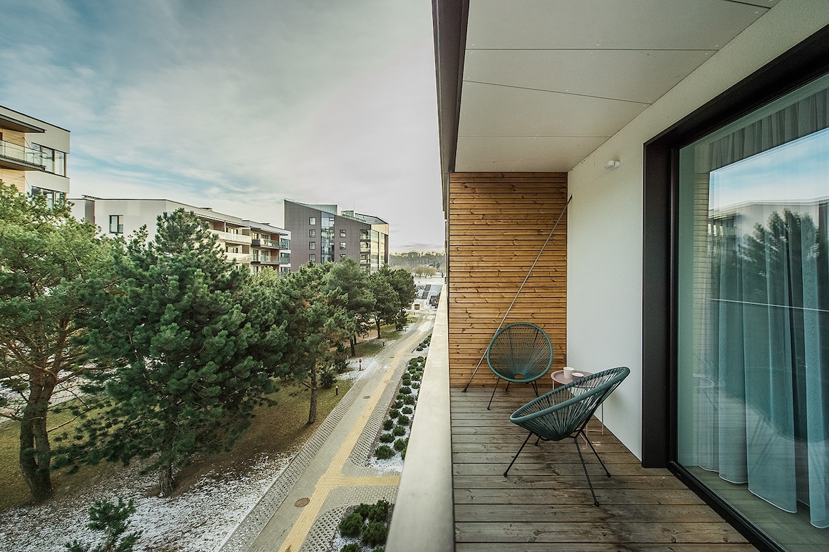 A private balcony is visible, featuring two comfortable chairs positioned against wooden paneling. Lush greenery and a pathway are seen below, with modern apartment buildings in the background. Natural light highlights the serene outdoor space, suitable for relaxation.