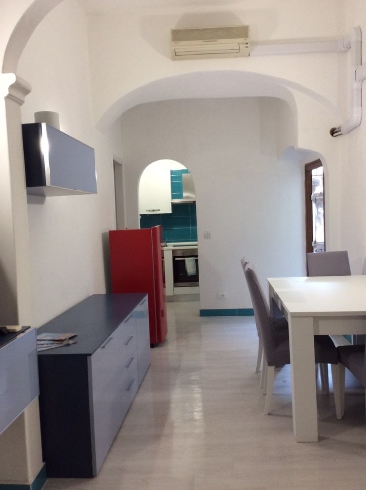 A modern kitchen area is visible, featuring a red refrigerator and a streamlined countertop under an arched ceiling. A dining table with four chairs occupies the space, and a glimpse of the kitchen cabinetry in blue is seen in the background.