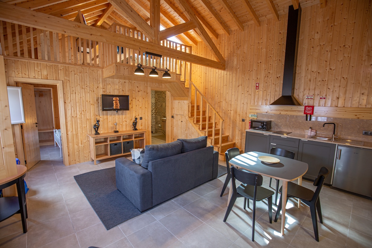 A spacious interior features wooden walls and beams, creating a warm ambiance. A comfortable sofa and dining table are situated in the open area, with natural light streaming through large windows. A staircase leads to a loft area, enhancing the sense of vertical space.