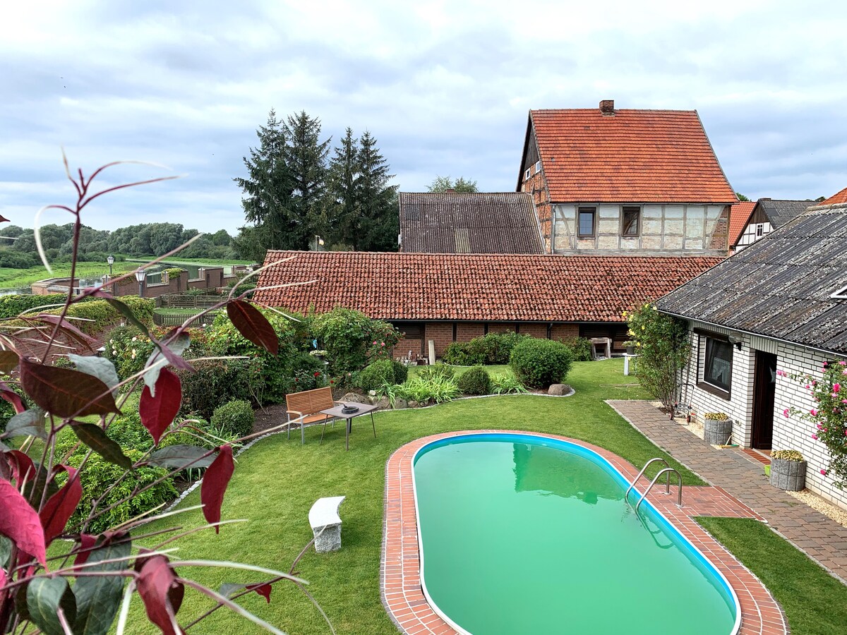 A spacious garden surrounds a rectangular swimming pool, inviting relaxation. The well-maintained outdoor area features green grass and wooden garden furniture. A rustic building with a red-tiled roof is visible in the background, complemented by a serene landscape of trees and fields.