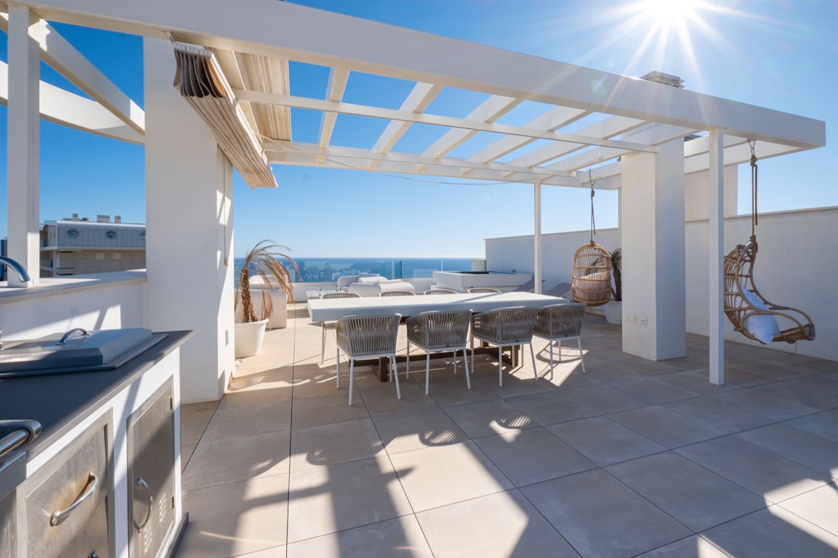 An open terrace features a spacious dining table surrounded by comfortable chairs. Two hanging chairs add a relaxed touch, and the outdoor kitchen is visible to the side. Bright sunlight casts shadows on the tiled floor, and panoramic views of the sea are seen in the background.