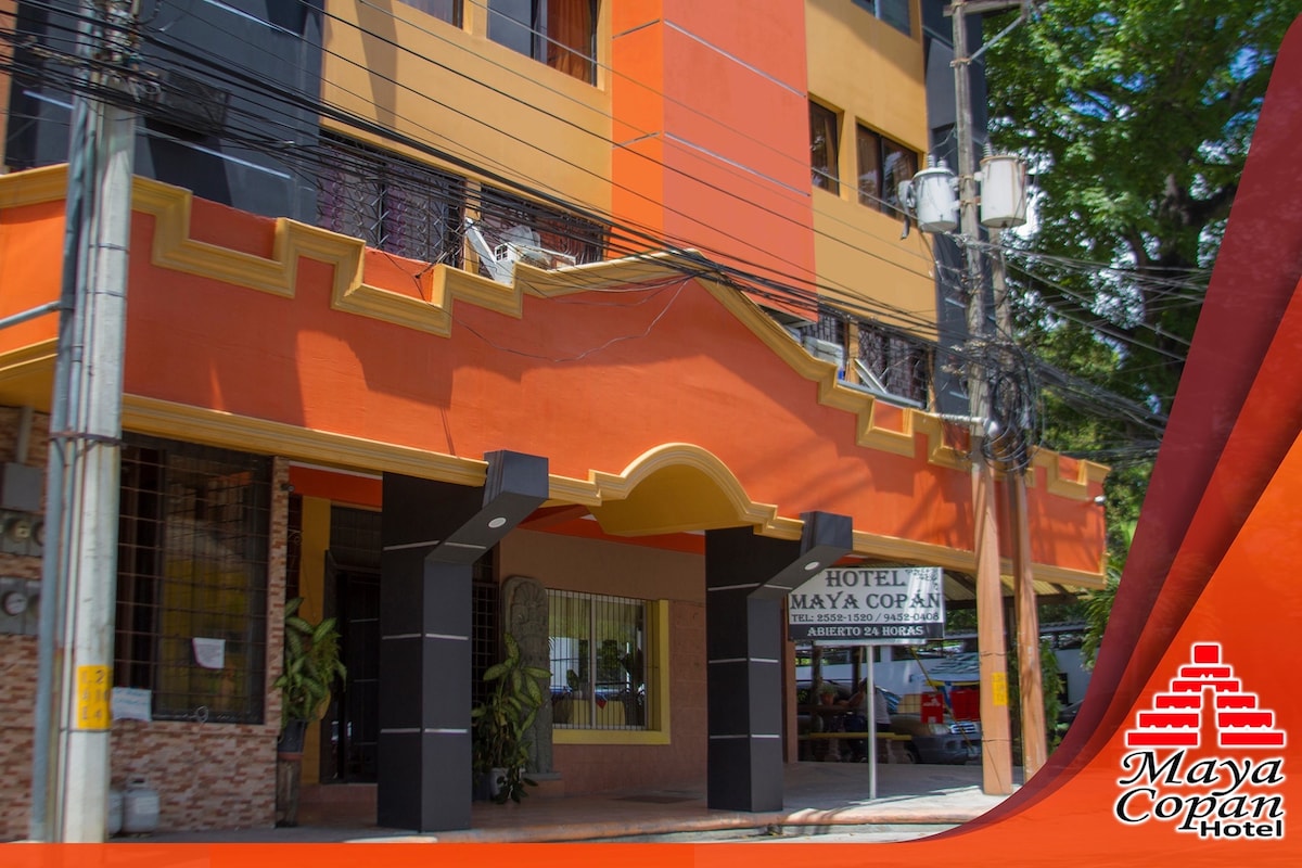 A vibrant building exterior is showcased, featuring bold orange and yellow accents. The entrance is marked by a decorative archway with signage indicating the hotel name. Lush greenery is visible near the entrance, contributing to the welcoming environment.