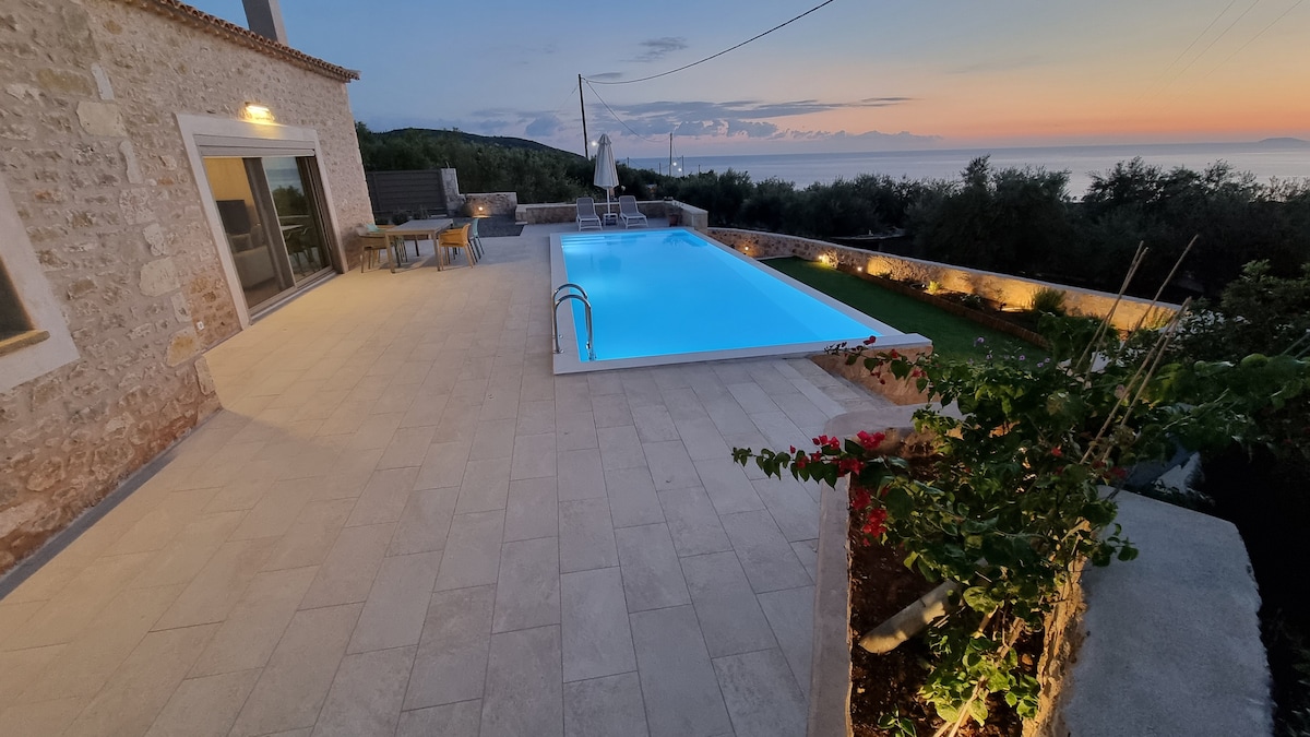 An outdoor area features a modern swimming pool surrounded by a stone terrace. Lounge chairs are positioned alongside the pool, while a dining set is seen nearby. Sunset colors create a serene backdrop with distant sea views and greenery framing the space.
