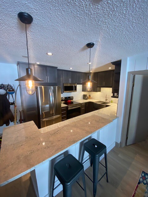 A modern kitchen is highlighted by dark cabinetry and an expansive countertop. Stainless steel appliances, including a refrigerator and oven, are visible. Two black stools are positioned at the bar area, and warm pendant lights hang above, creating a functional and inviting space.
