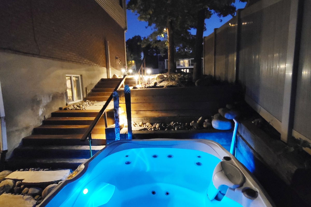 An outdoor hot tub is illuminated in soft blue light, creating a serene atmosphere. Surrounding landscaping features stone pathways and wooden steps, leading to a higher area with additional greenery. The scene is set during twilight, enhancing the tranquil environment.