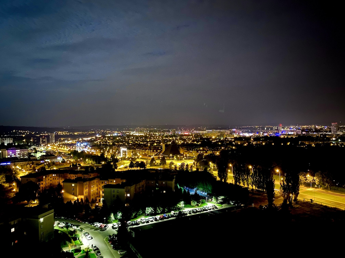 A nighttime view offers a sweeping panorama of a city illuminated by streetlights and building facades. Various patterns of light create dynamic visuals across the landscape, with a dark sky above. The scene conveys the vibrant atmosphere of urban living after sunset.