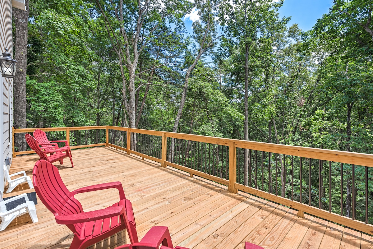 A wooden deck extends into a natural setting, featuring several red adirondack chairs. Lush green trees surround the area, creating a sense of privacy and tranquility. The sun filters through the leaves, enhancing the inviting outdoor space.