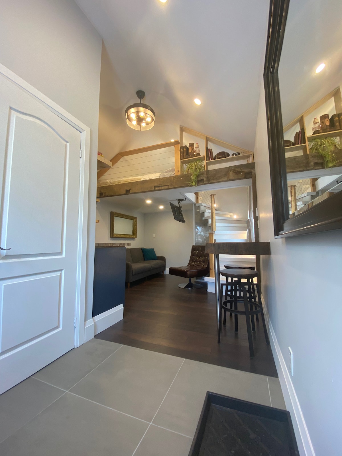 An entryway features sleek tiled flooring leading into the living area, where a pull-out couch is positioned. A staircase with wooden detailing ascends to the lofted sleeping area. Natural light is provided by overhead fixtures, enhancing the open layout and modern design.