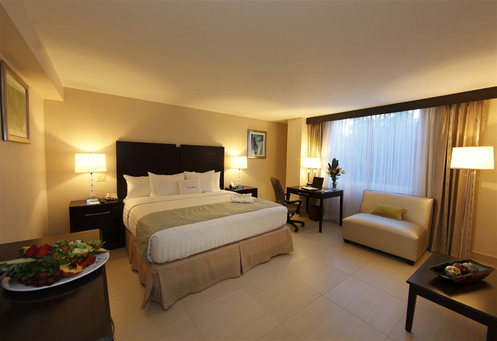 A spacious room features a large bed with crisp white linens and a decorative headboard. Two bedside lamps provide soft lighting, while a desk and chair are positioned by a window, complemented by a comfortable seating area. Fresh fruit is displayed on a nearby table.