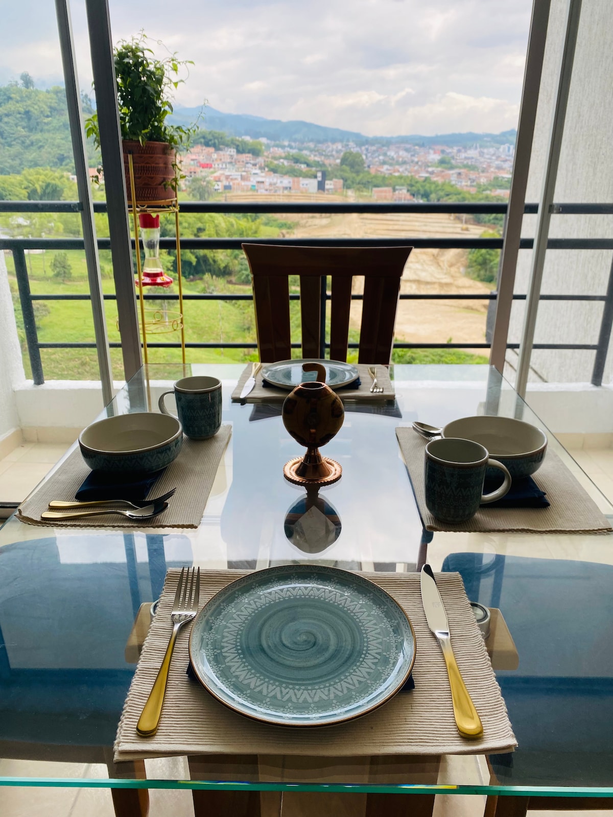 A dining table is set for four, featuring polished utensils and plates placed atop neutral placemats. The view beyond showcases green hills and cityscapes through large glass windows. A hummingbird feeder hangs nearby, enhancing the serene dining atmosphere.
