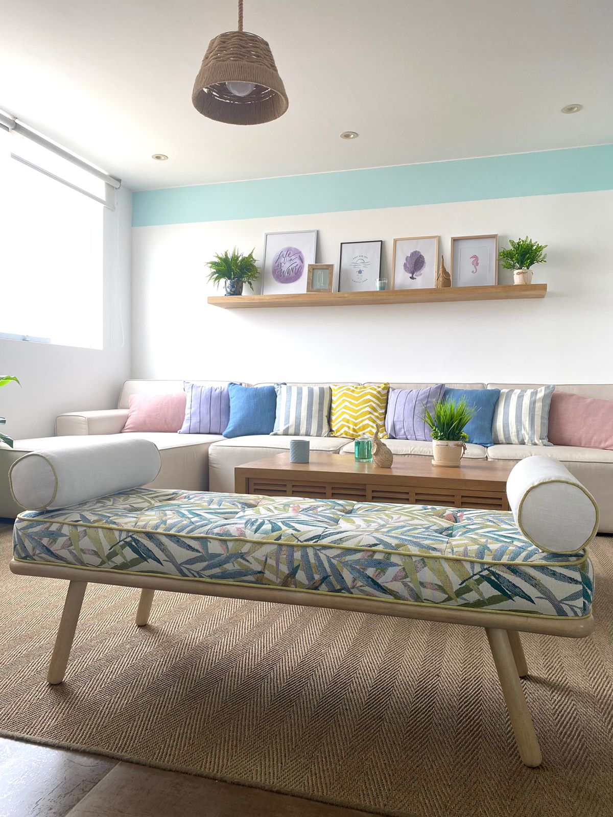 A bright seating area features a patterned bench with rounded armrests, positioned across from a light-colored sofa adorned with pastel pillows. A wooden coffee table is centered in the space, complemented by wall-mounted shelves displaying framed art and potted plants.