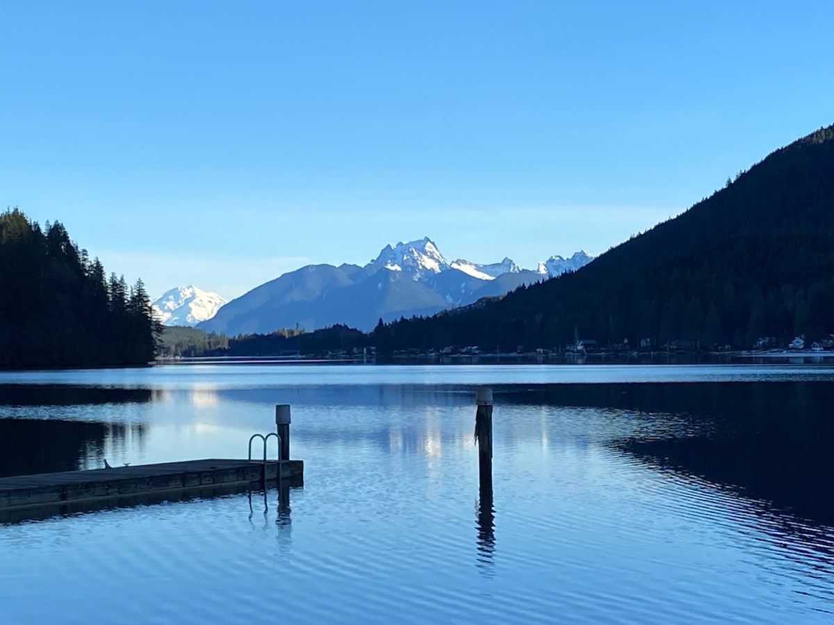 A serene view of Lake Cavanaugh is presented, with calm waters reflecting surrounding mountains. The scene includes a wooden dock extending into the lake, along with forested hillsides framing the backdrop. Snow-capped peaks rise prominently against a clear blue sky.