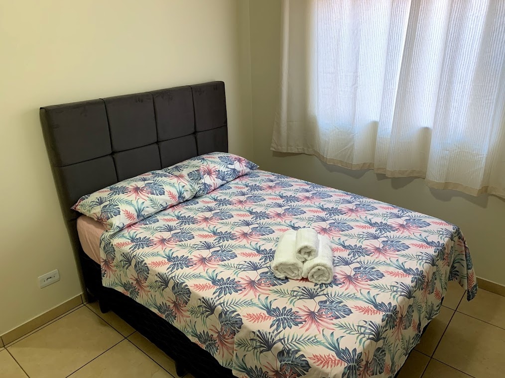 A cozy bedroom features a spacious bed with a patterned coverlet, complimented by two neatly rolled towels on top. Natural light filters through a sheer window treatment, creating a comfortable and inviting space.